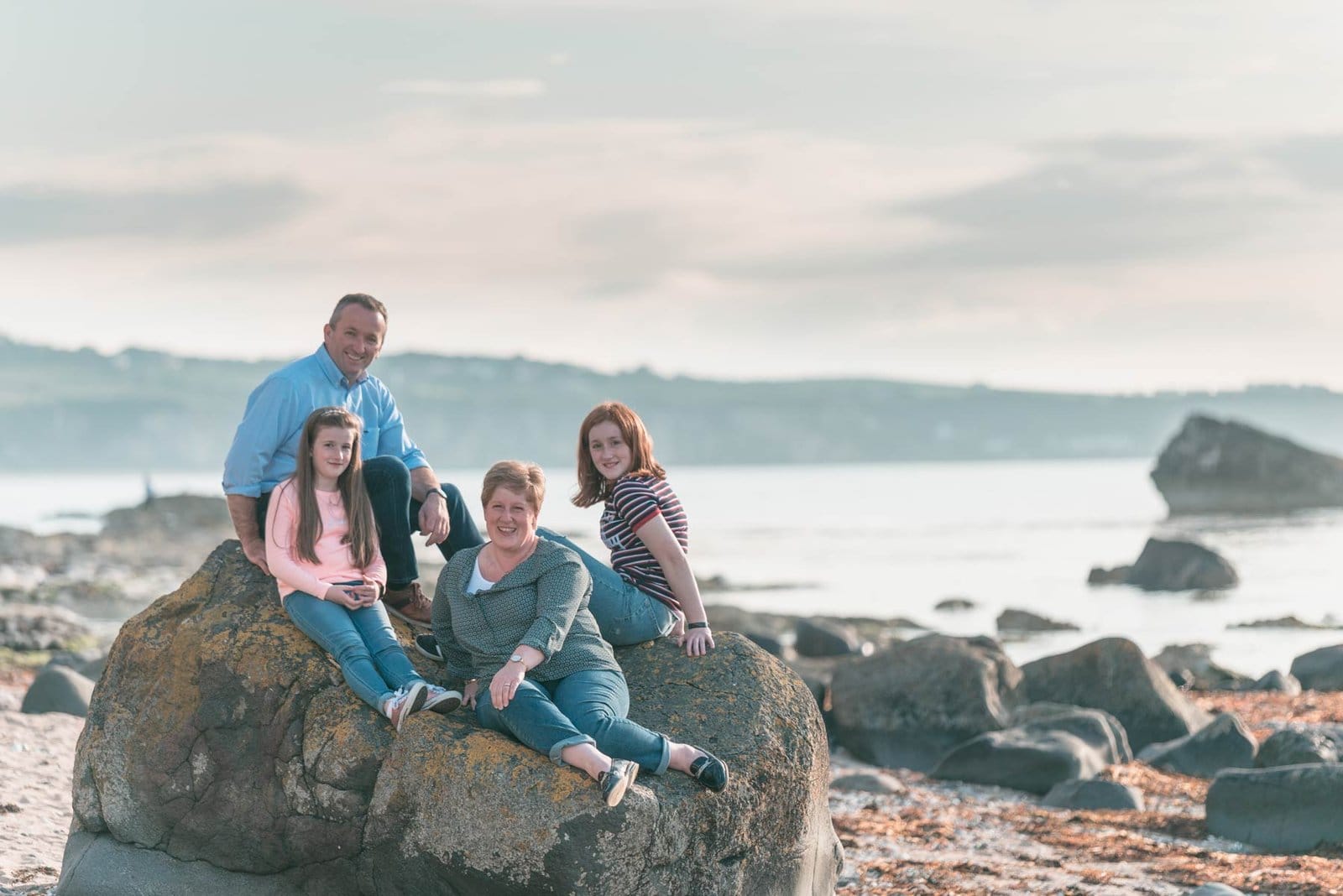 A Northern Ireland family sits on rocks on the beach. Northern Ireland Family Photographer
